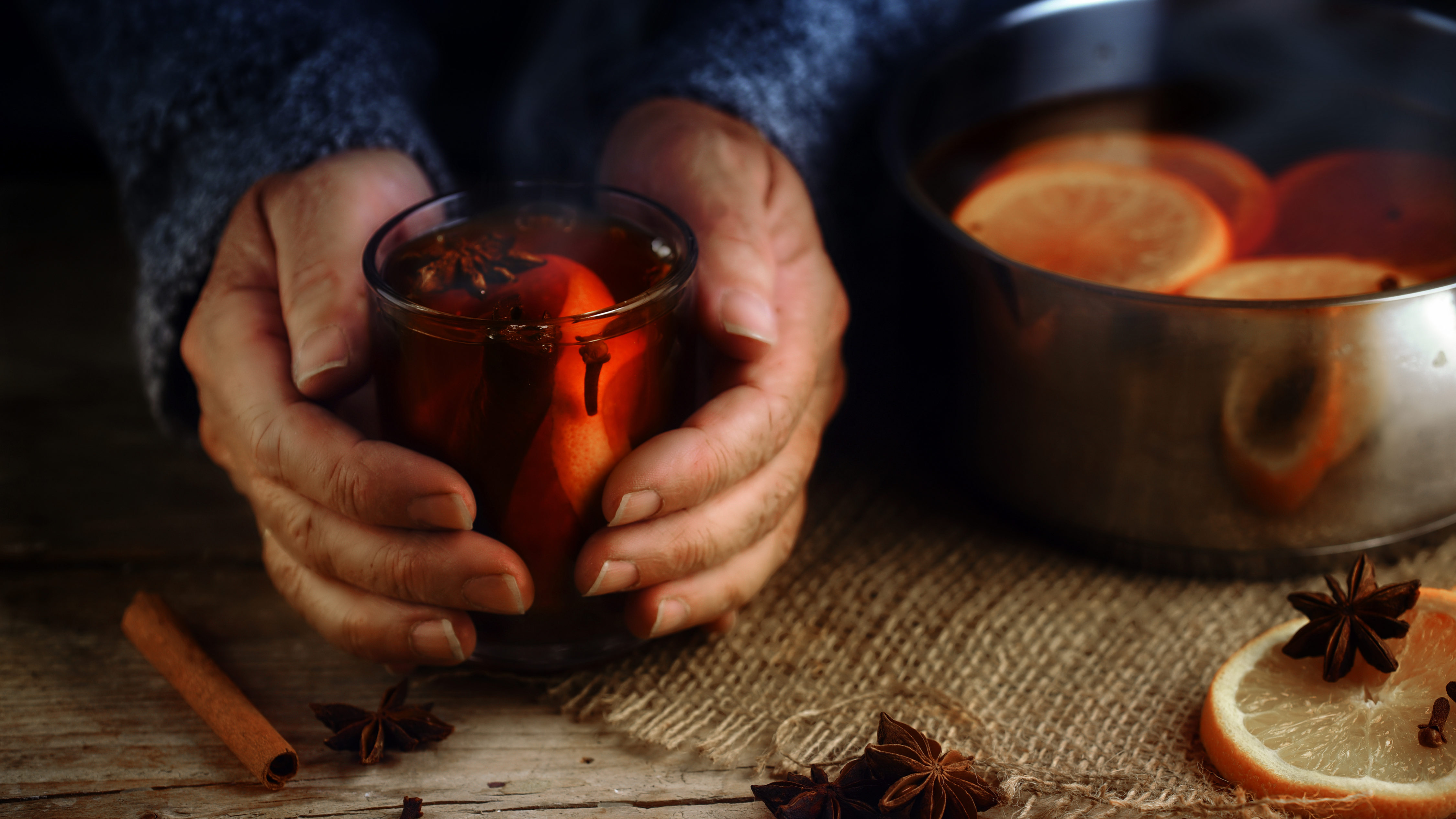 Older Female Hands Holding A Glass Mug With Hot Mulled Wine Next To The Steaming Cooking Pot, Christmas Ingredients, Orange Slices, Cinnamon Sticks, Star Anise And Cloves, A Warming Home Scene On A Rustic Wooden Table