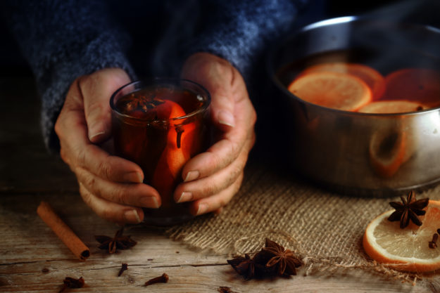 Older Female Hands Holding A Glass Mug With Hot Mulled Wine Next To The Steaming Cooking Pot, Christmas Ingredients, Orange Slices, Cinnamon Sticks, Star Anise And Cloves, A Warming Home Scene On A Rustic Wooden Table